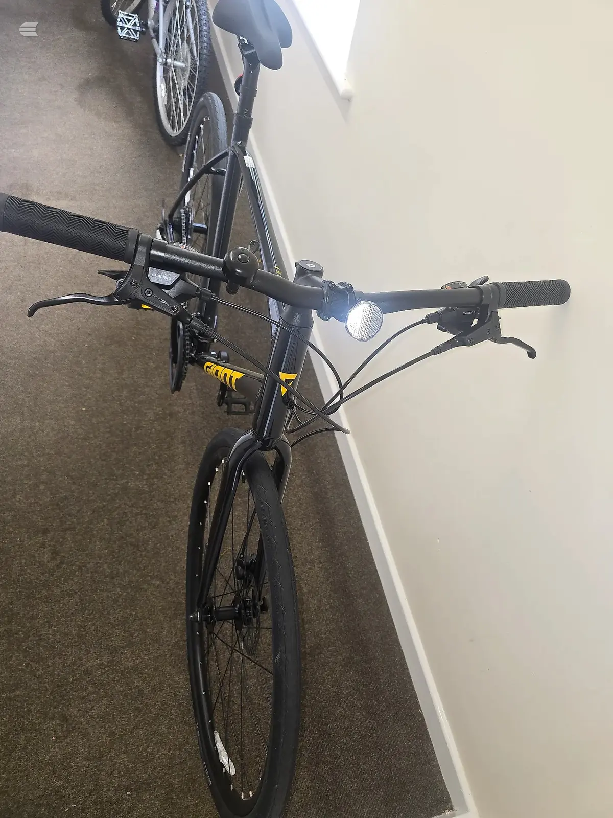 Black hybrid bike with yellow frame accents, front-facing view indoors against white and grey walls, headlight visible on handlebars.