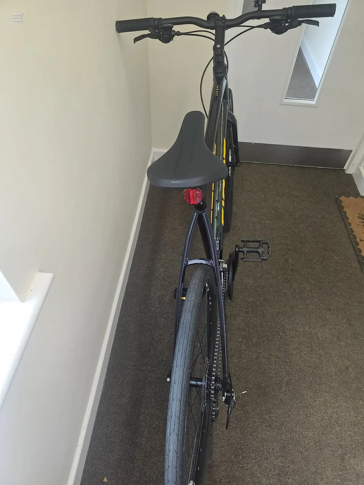 Black commuter bike with yellow accents, viewed from the right side in an indoor hallway. Bike is stationary on dark carpet against white walls.