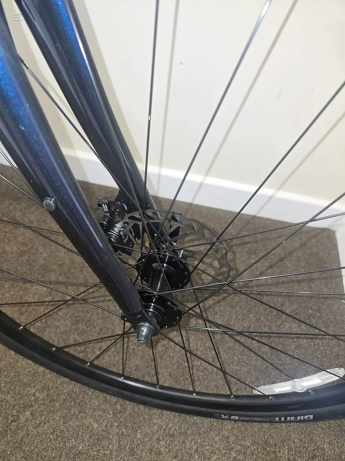 Front wheel of a dark blue hybrid or commuter bike with disc brakes, close-up view indoors on carpet, spokes and hub in focus.