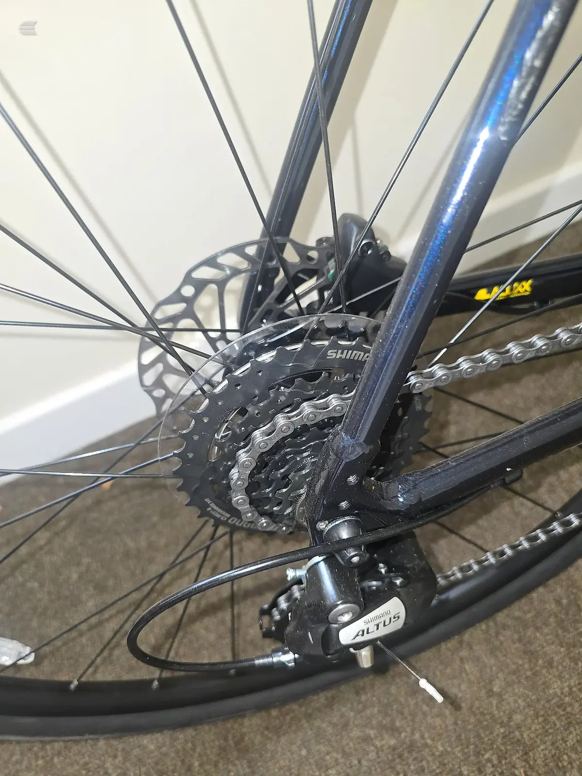Close-up of rear drivetrain on blue mountain bike, showing Shimano cassette, chain, and derailleur, drive side, indoors on carpet.