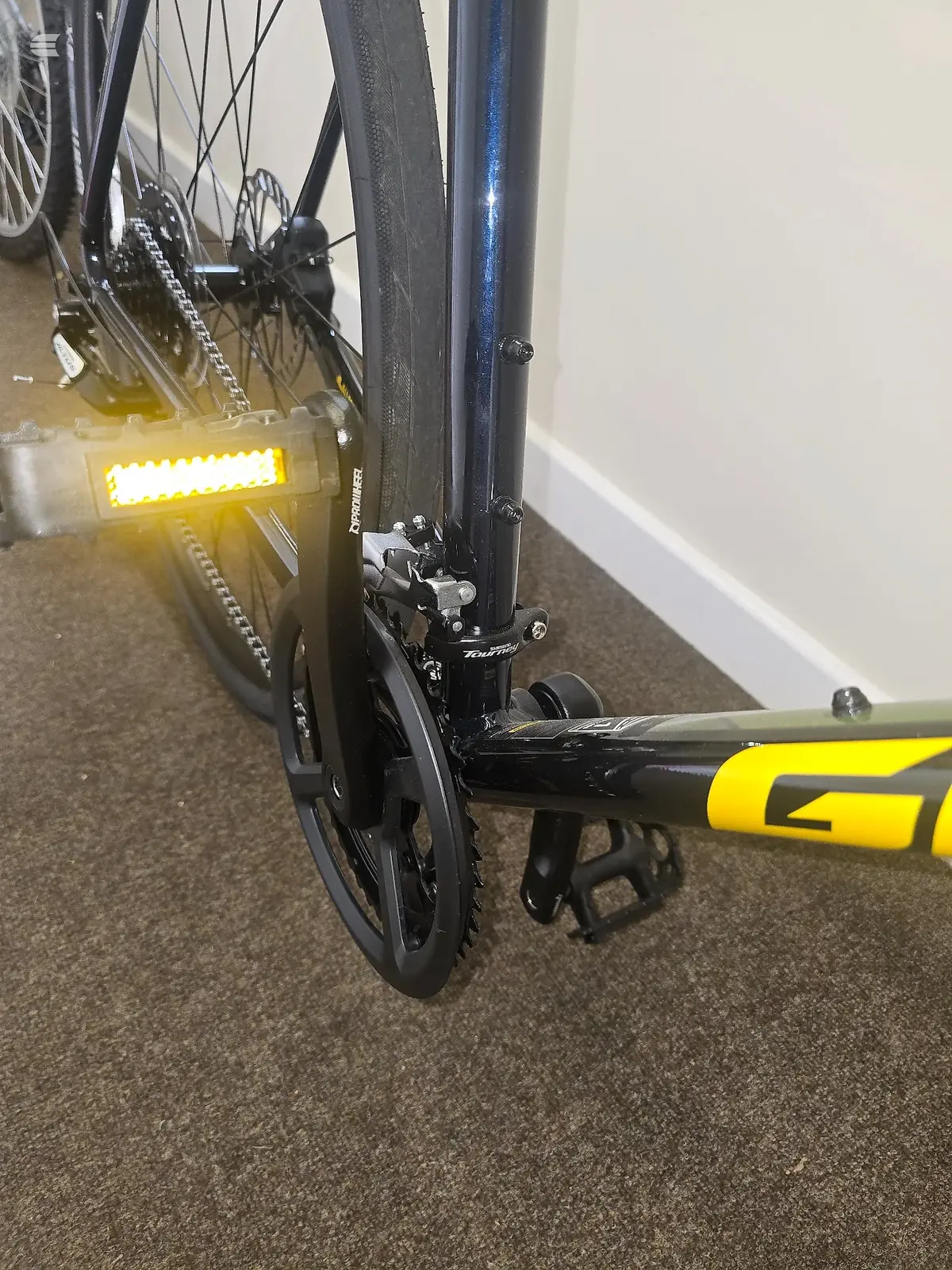 Close-up of rear derailleur and cassette on a blue and yellow bike, drive side, indoors on grey carpet against white wall.