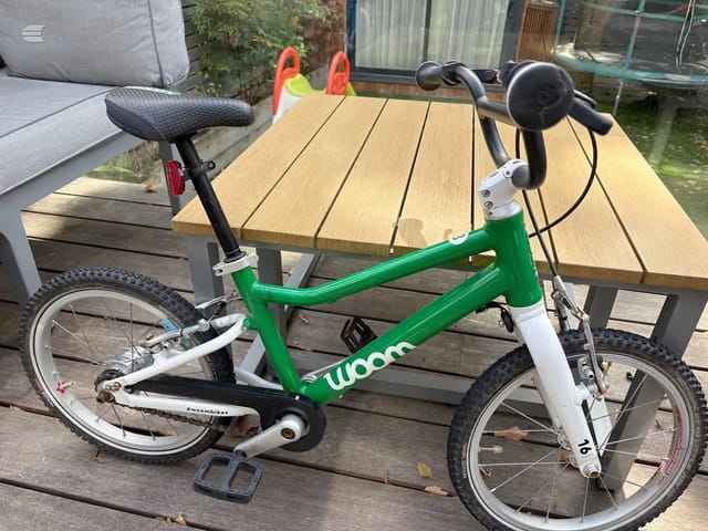 Green and white Woom children's bike on wooden deck, drive side, garden setting with table and trampoline behind.