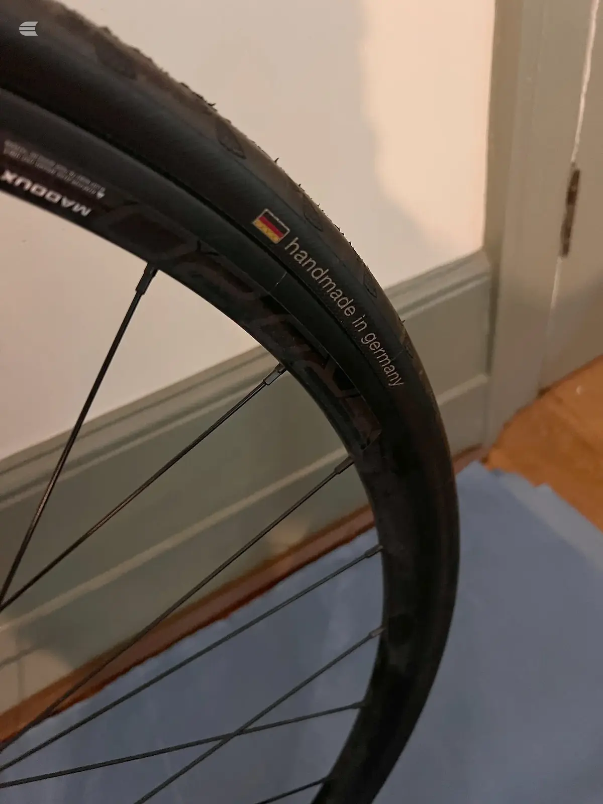Close-up of black bicycle tyre and rim with German 'handmade in Germany' text visible, angled against white door frame and wooden floor.