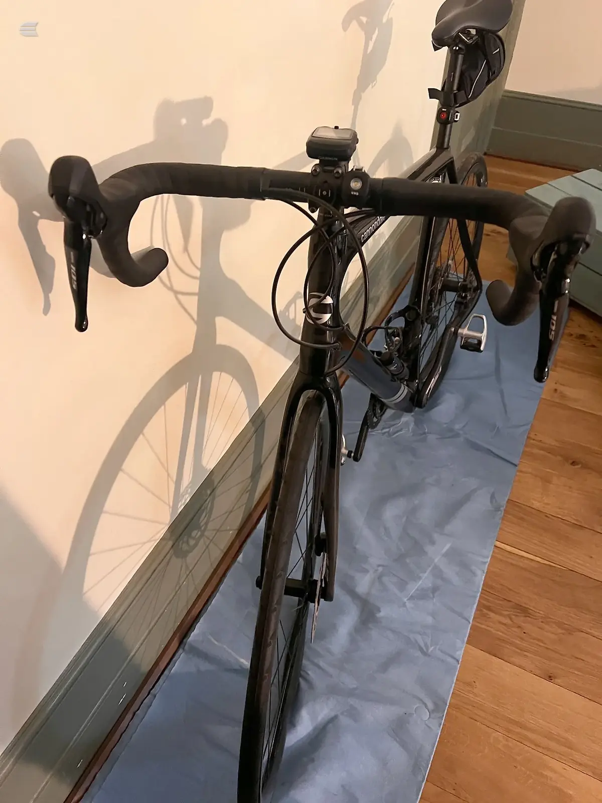 Black drop-bar road bike on blue mat indoors, drive side, against white wall with strong shadow cast.