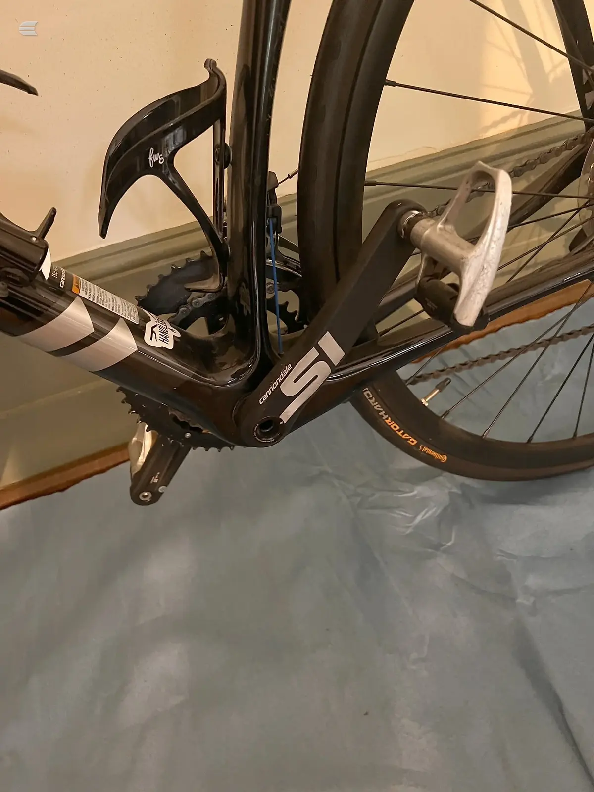Black Cannondale road bike, drive-side view of bottom bracket, crankset, and pedals on concrete floor indoors.