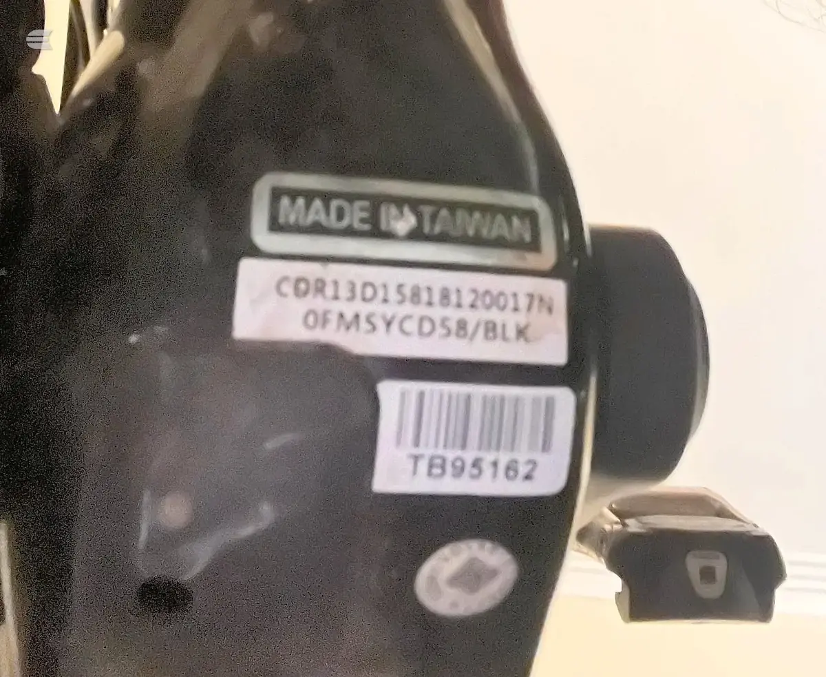 Close-up of black bicycle component with Taiwan manufacturing label and product code stickers, against cream background.