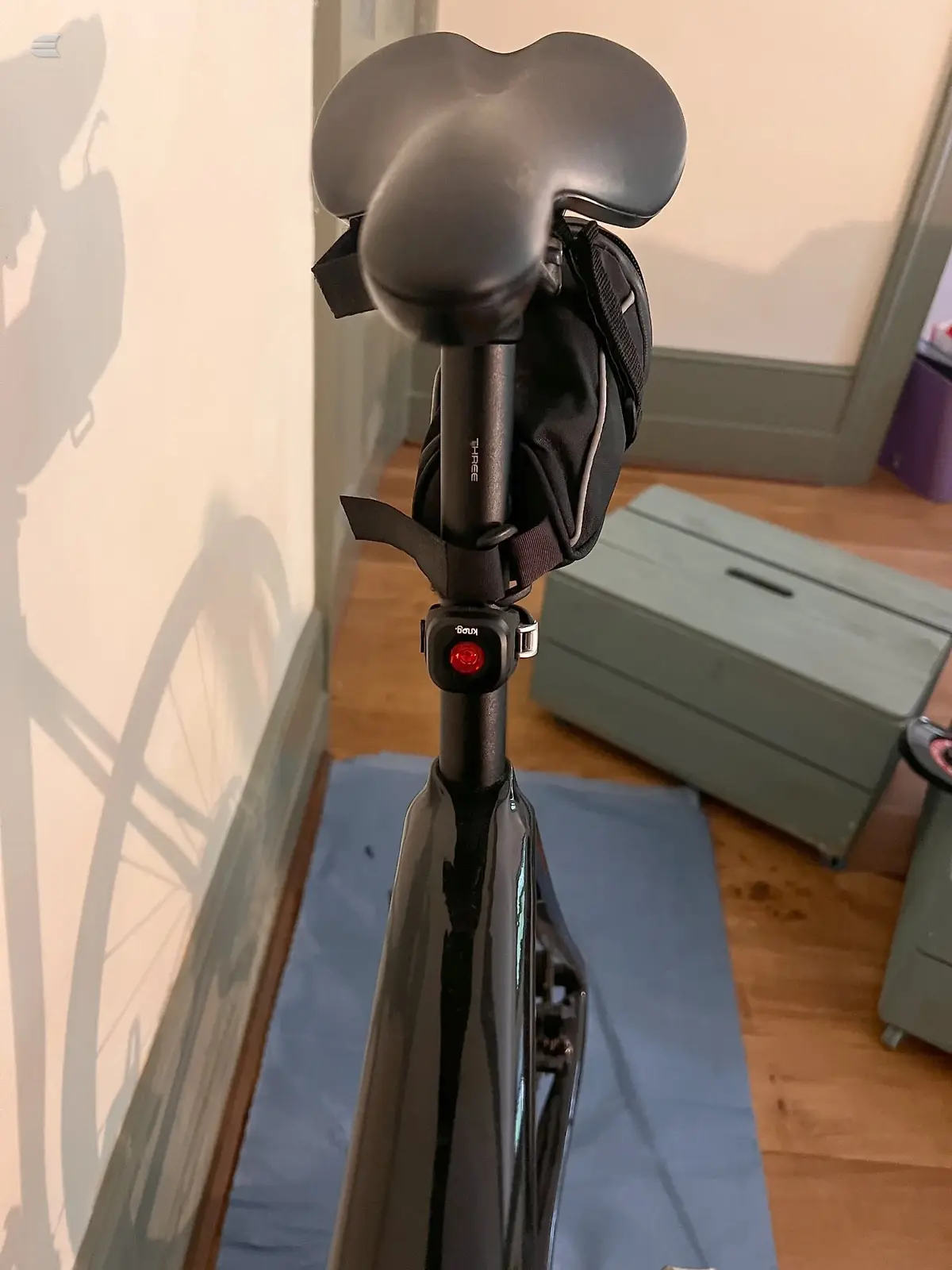 Black stationary bike on blue mat indoors, non-drive side view, against white wall with natural light casting shadow.
