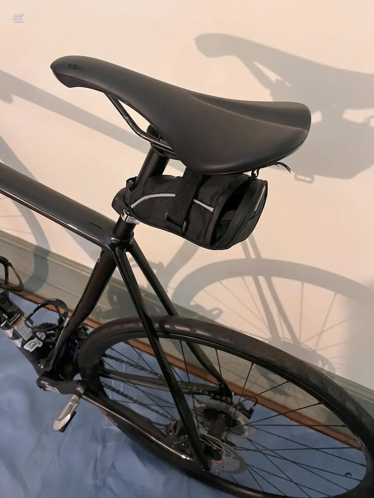 Black hybrid or commuter bike on a stand, drive side, against a beige wall with strong shadow cast by the bicycle frame and wheels.