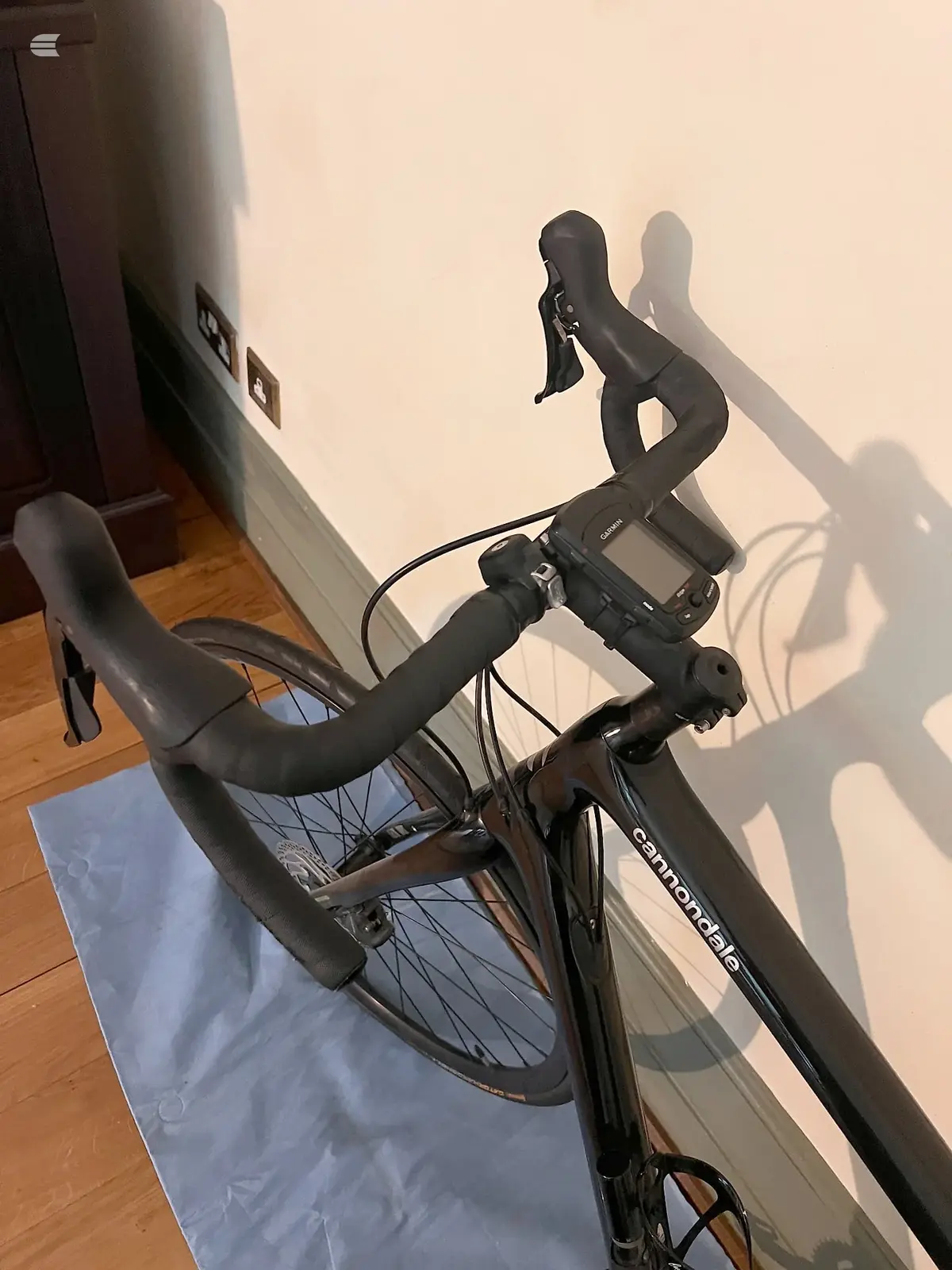 Black Cannondale road bike with drop bars on a blue mat indoors, non-drive side, casting a shadow on a cream wall.