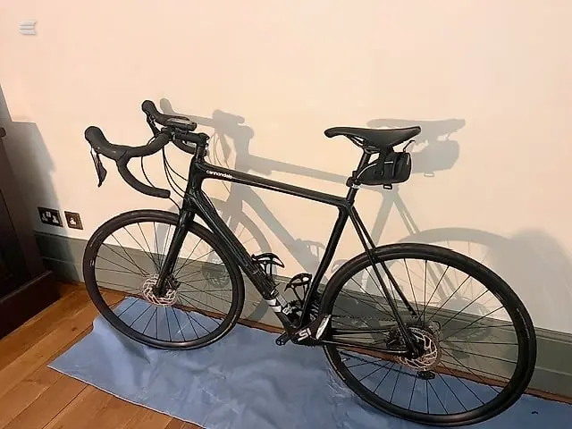 Black road bike on blue mat indoors, drive side, against white wall with strong shadow cast by window light.