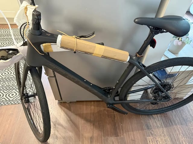 Black gravel bike indoors on wooden floor, non-drive side, with tan handlebar tape and red rear light visible.