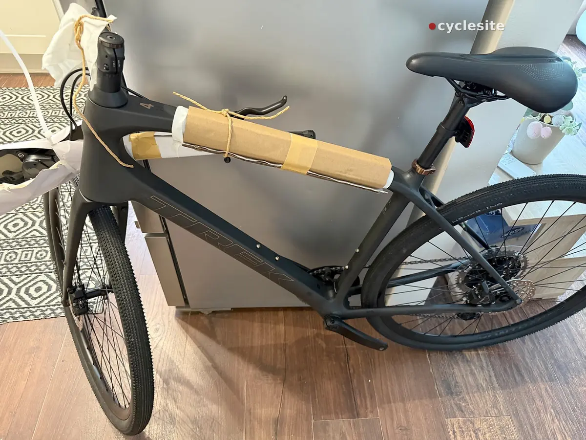 Black gravel bike indoors on wooden floor, non-drive side, with tan handlebar tape and red rear light visible.