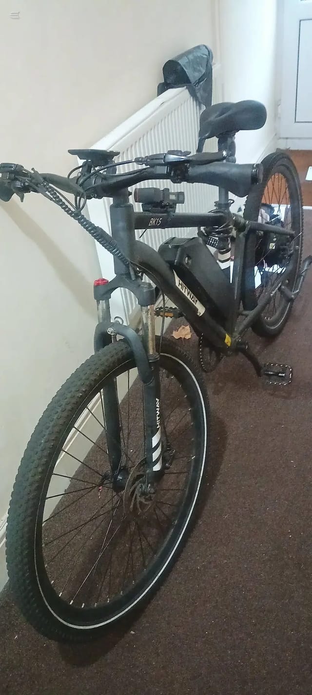 Black hardtail mountain bike on brown carpet indoors, non-drive side, leaning against white radiator and wall.