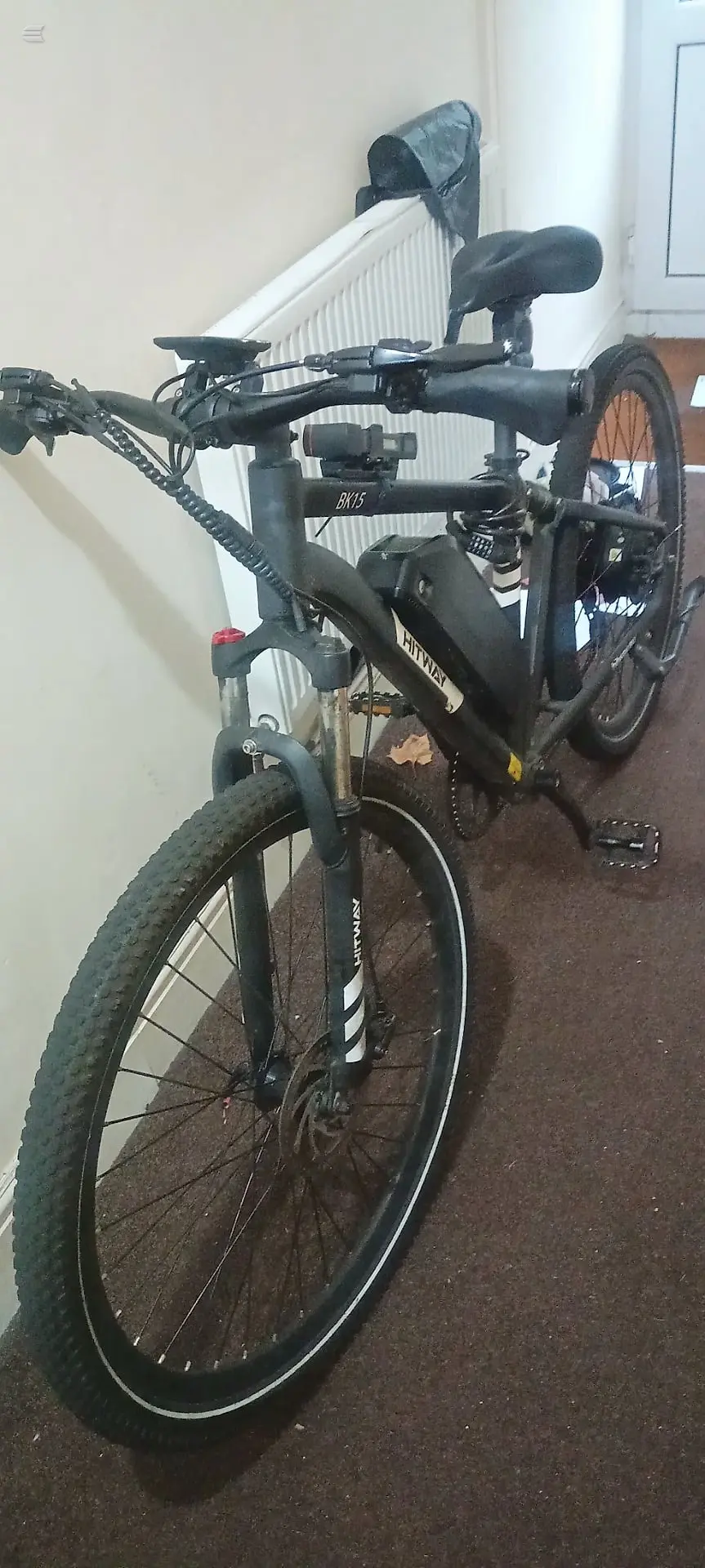 Black hardtail mountain bike on brown carpet indoors, non-drive side, leaning against white radiator and wall.