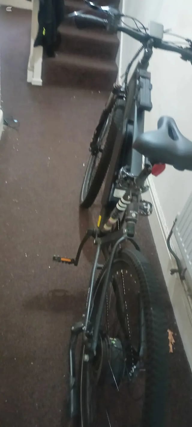 Black hybrid or commuter bike indoors against a brown wall, non-drive side, tilted at an angle with saddle and handlebars visible.