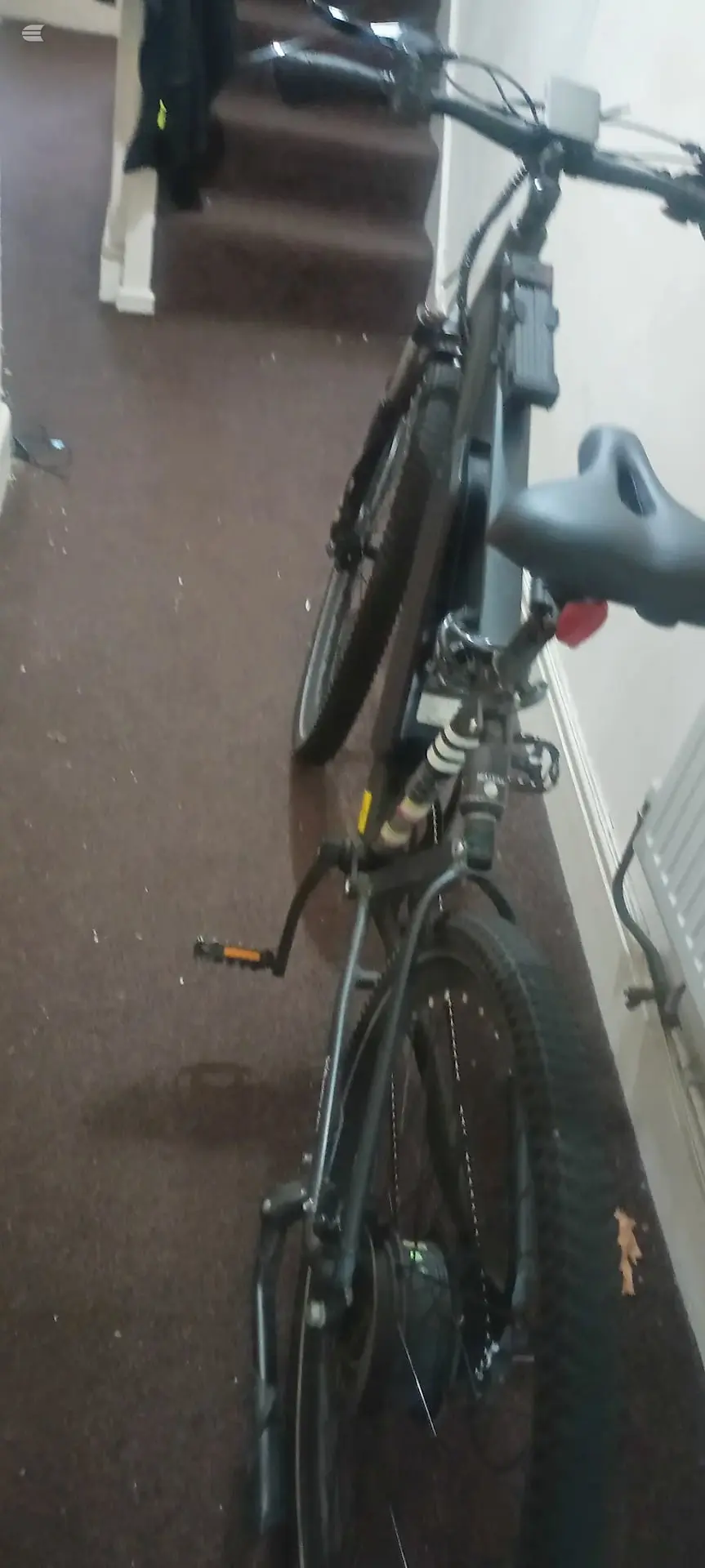 Black hybrid or commuter bike indoors against a brown wall, non-drive side, tilted at an angle with saddle and handlebars visible.