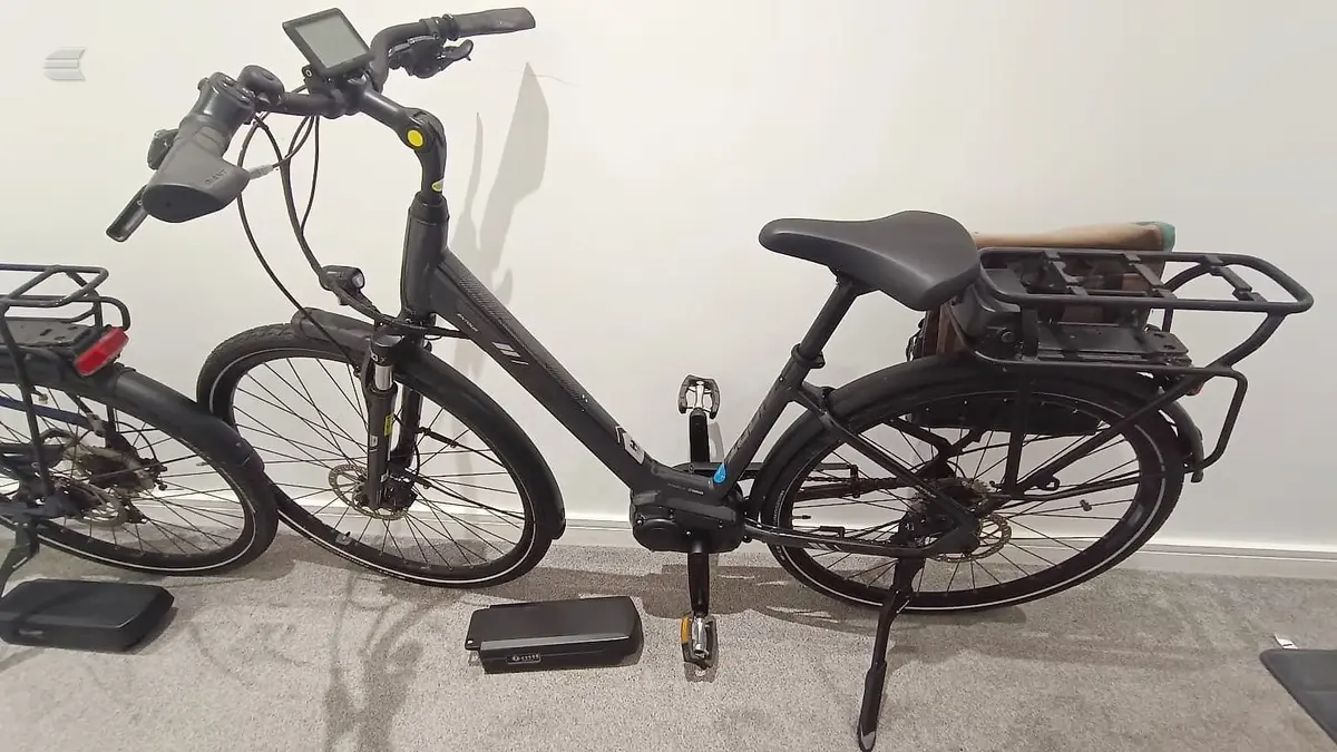 Two black electric cargo bikes indoors against a white wall, non-drive side view. Left bike has front basket and digital display; right bike has rear cargo