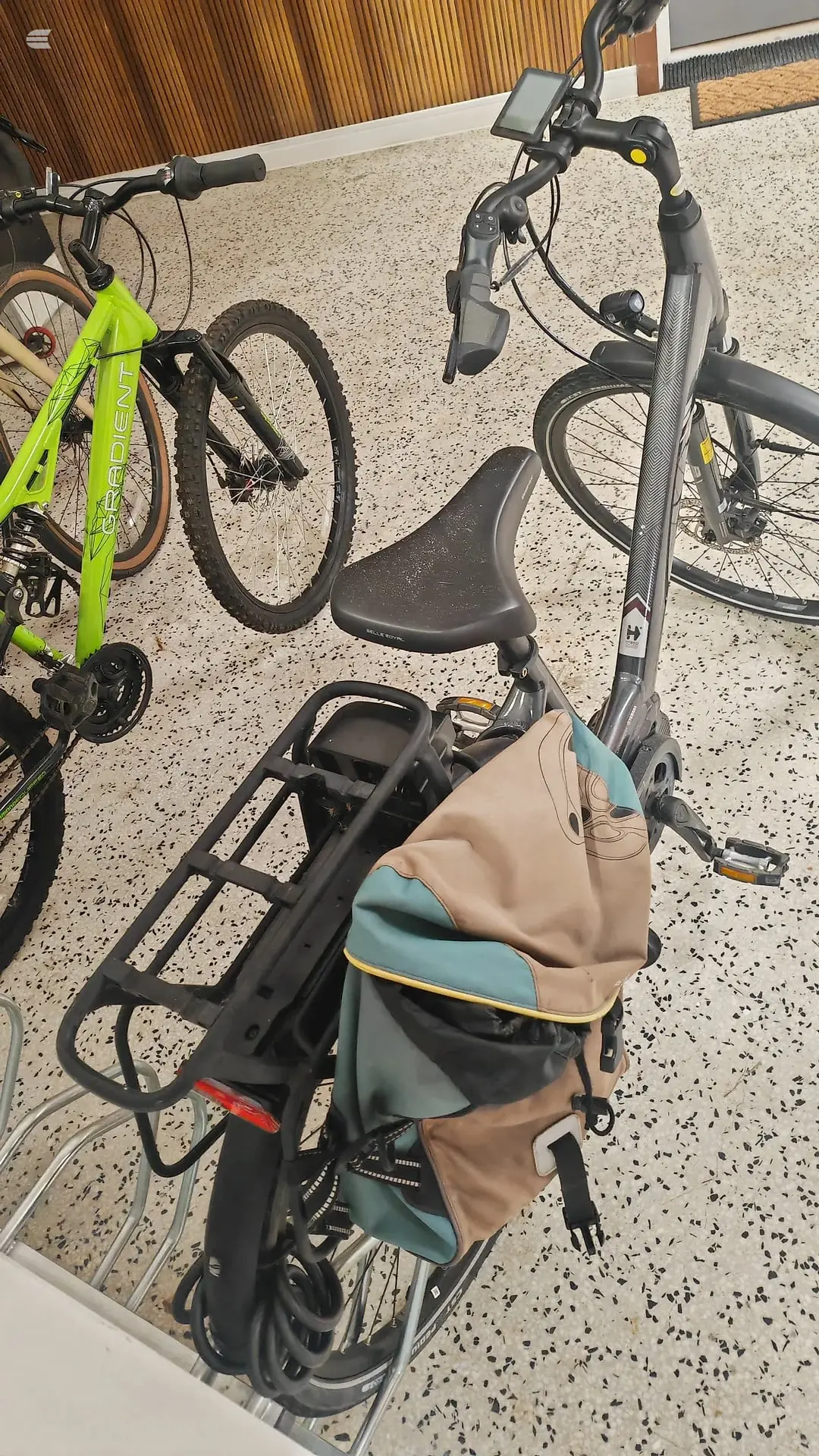 Grey electric bike with integrated display and front suspension, parked indoors on speckled floor next to a neon-yellow hardtail mountain bike, three-quarter