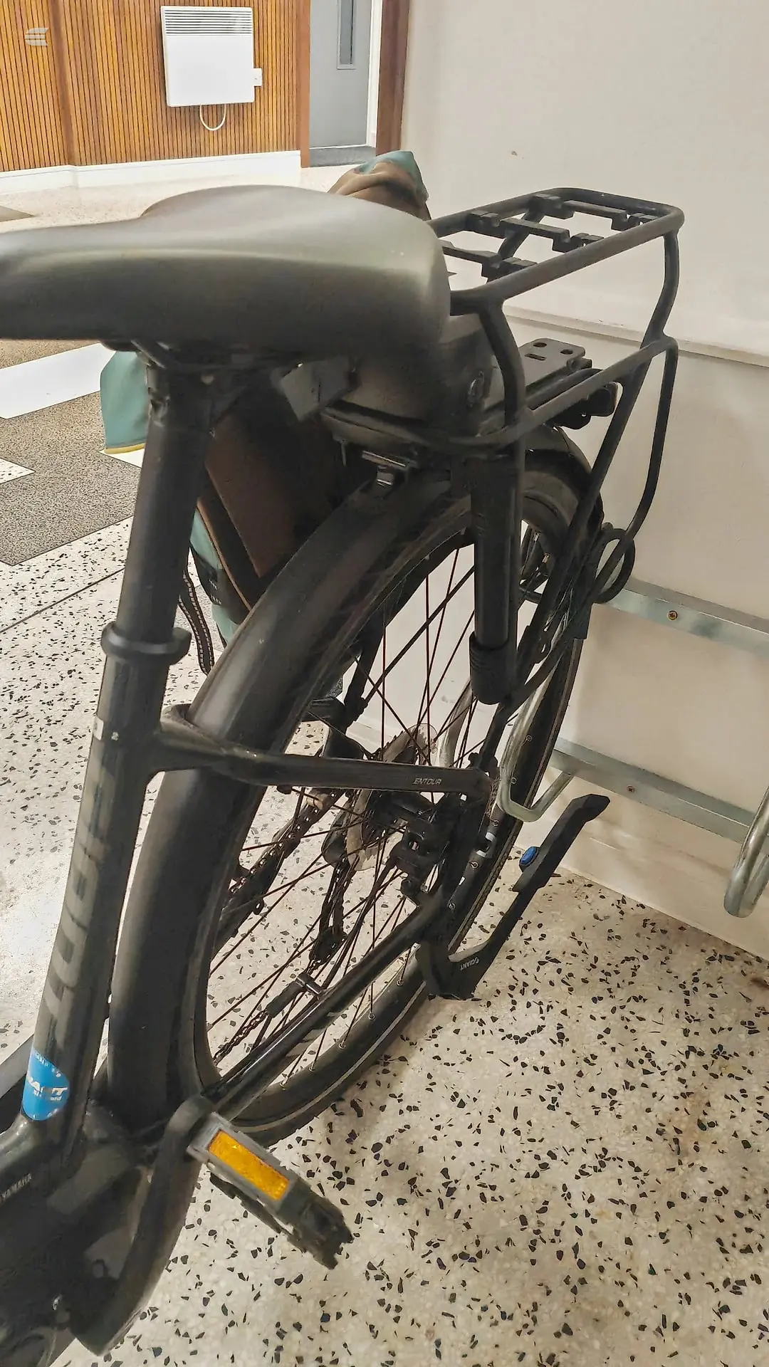 Black utility bike with rear cargo rack, non-drive side, indoors on speckled floor against white wall.