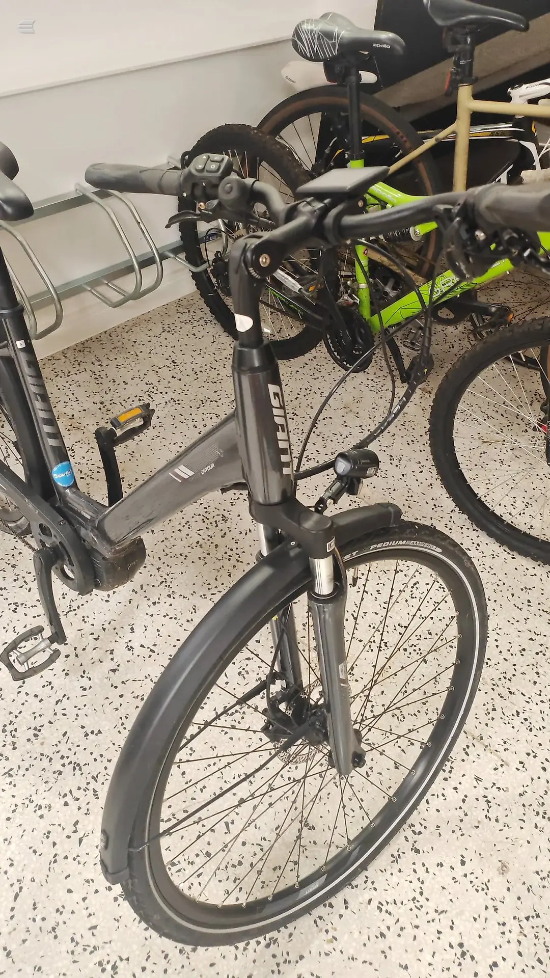 Grey commuter bike with mudguards on concrete floor, three-quarter view from drive side, parked indoors next to a green bike and bike rack.