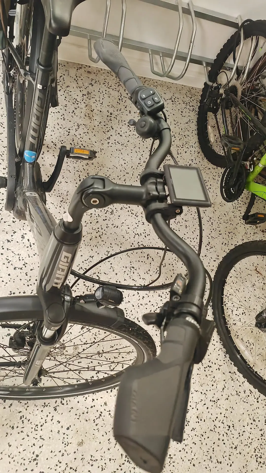 E-bike handlebars and cockpit viewed from above on speckled concrete floor, showing display screen, brake levers, and shifter controls. Multiple bikes parked