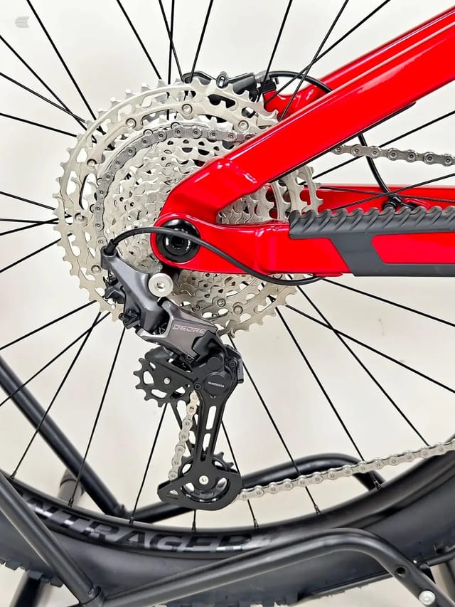 Red mountain bike rear drivetrain close-up showing Shimano Deore derailleur, cassette, and chain against white background, drive side.