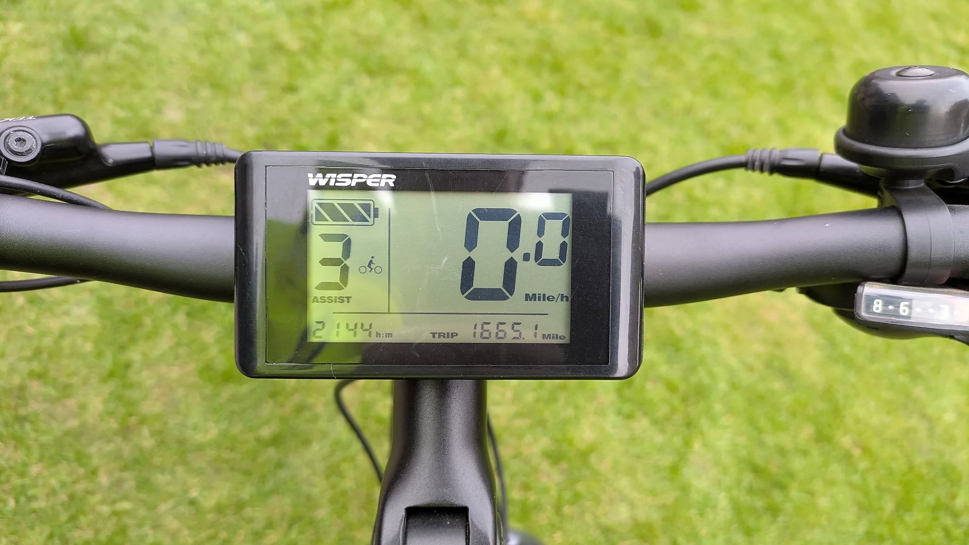 Wisper electric bike display console mounted on handlebars, showing speed of 20.0 mph, trip distance 1865.1 miles, and battery level, against green grass