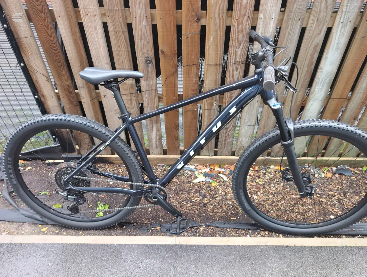 Black hardtail mountain bike on gravel ground, drive side, against wooden fence with autumn leaves.
