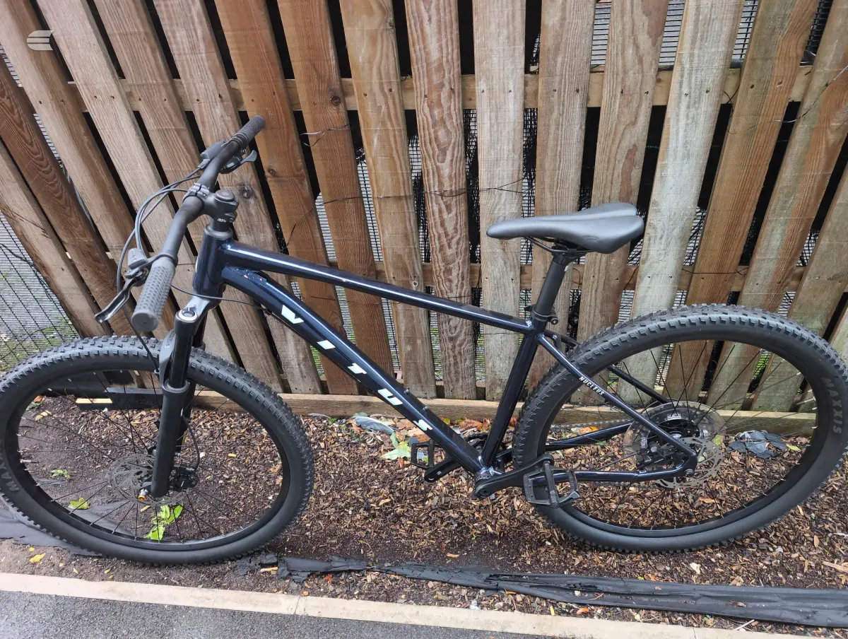 Black hardtail mountain bike on bark mulch, drive side, against wooden fence.