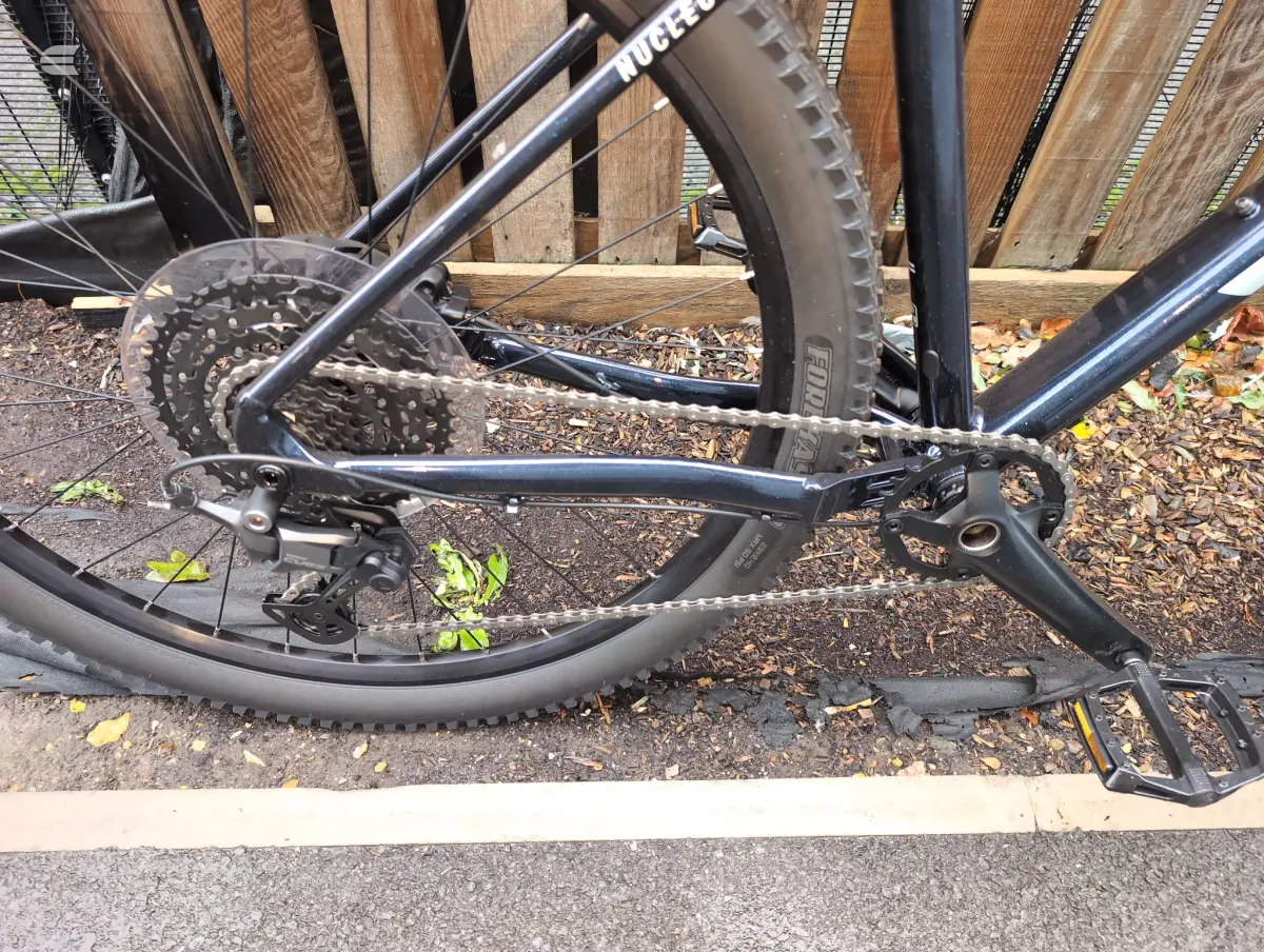 Black gravel bike, drive side, close-up of rear wheel and drivetrain on wood deck with autumn leaves, against wooden fence.