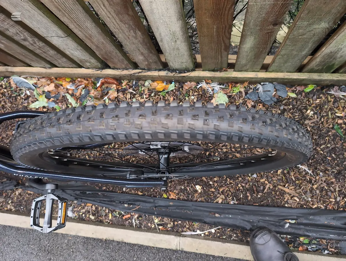 Mountain bike front wheel with knobby tread tyre, mounted on black fork, non-drive side view, against wooden fence and leaf litter.
