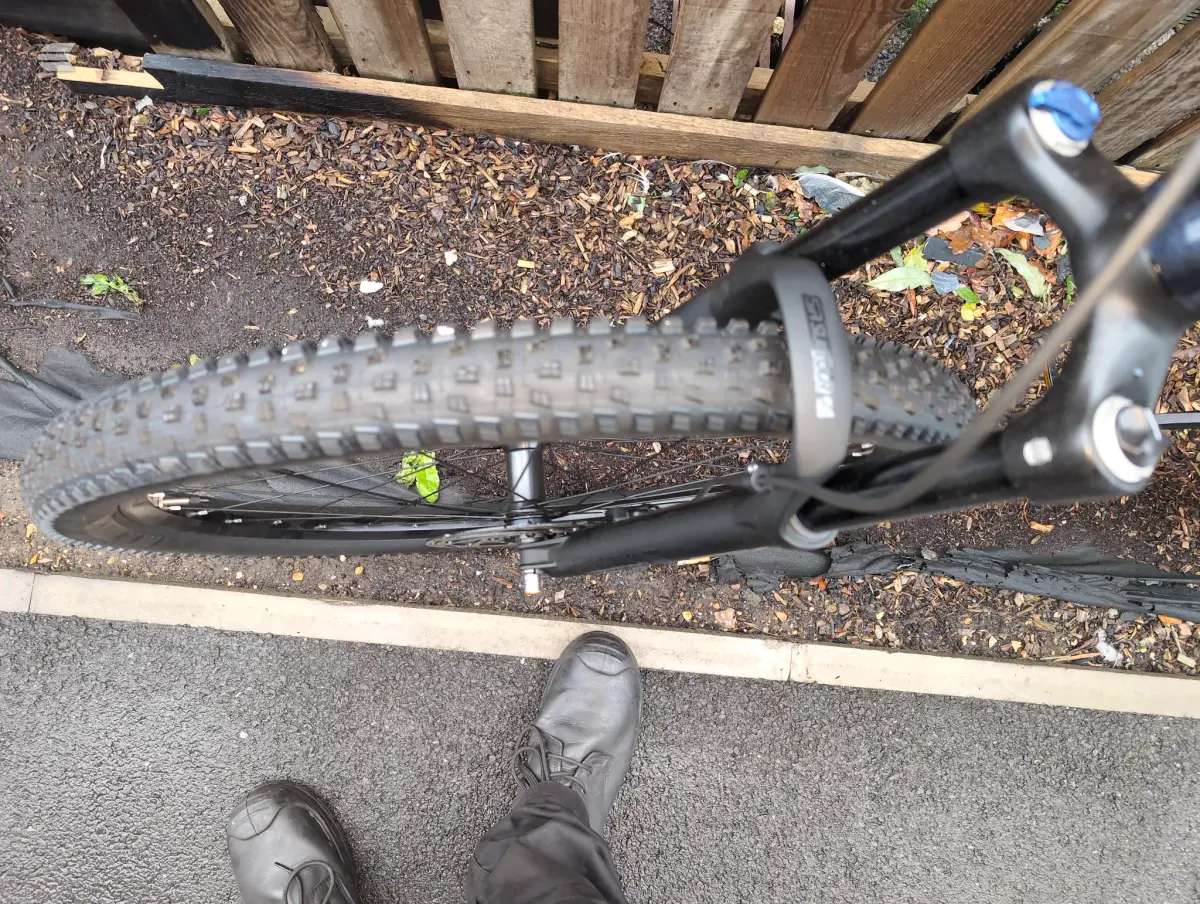 Black hardtail mountain bike on wood chips and gravel, overhead view, drive side, with rider's boots visible at bottom edge.