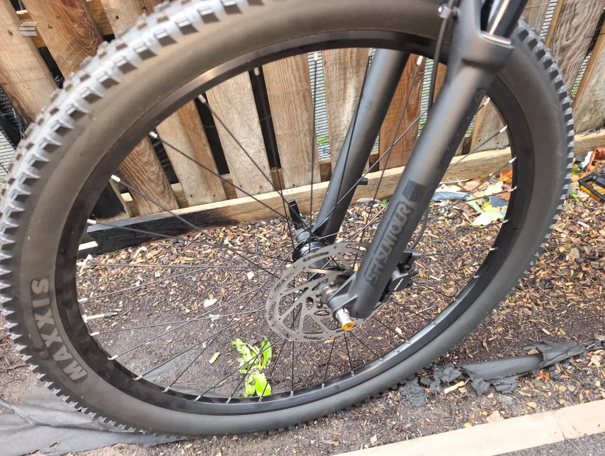 Front wheel of a grey hardtail mountain bike, close-up on drive side, against wooden fence and mulch ground.