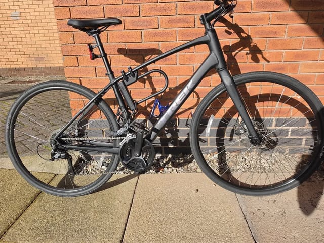 Black gravel bike with disc brakes on concrete paving against red brick wall, drive side, full frame visible with shadow cast on wall.