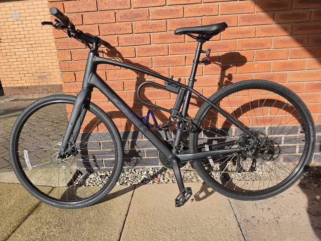 Black hybrid bike on gravel against red brick wall, drive side, in bright sunlight.