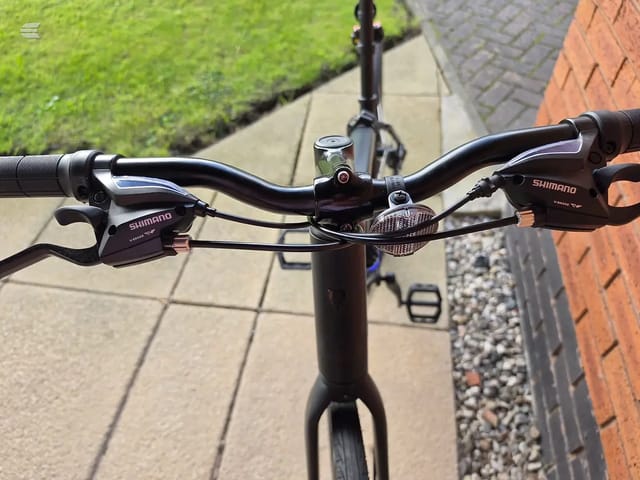 Close-up of Shimano brake levers and handlebars on a hybrid or commuter bike, viewed from above on a paved driveway with brick wall and grass visible.