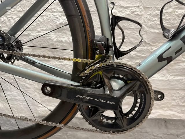 Close-up of Shimano Ultegra crankset and chain on light blue road bike frame, drive side, against white textured wall.