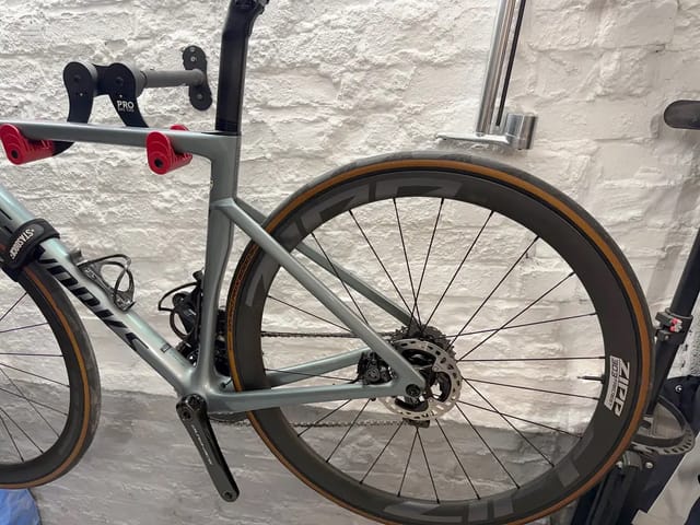 Grey gravel bike on wall-mounted stand, drive side, against white brick wall. Brown-wall tyres, disc brakes, drop bars with red bar-end plugs visible.