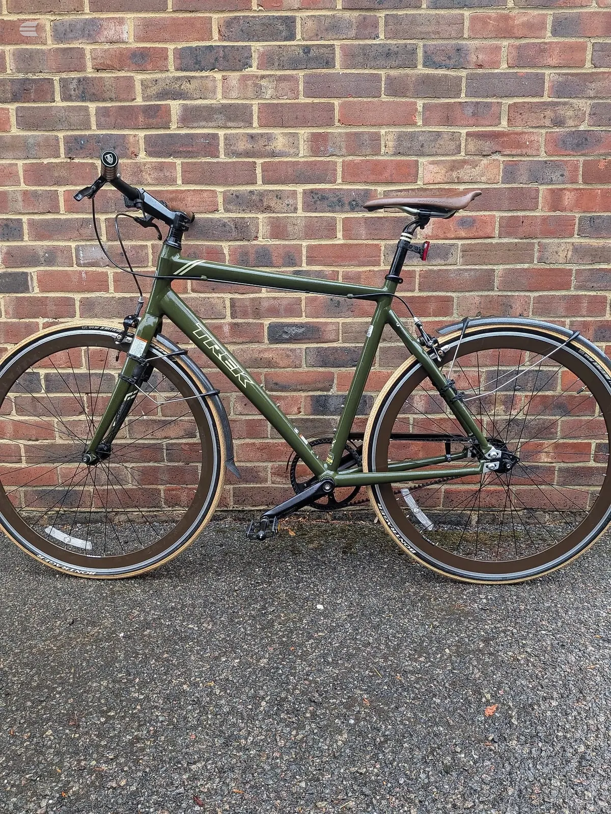 Olive green hybrid bike on asphalt, drive side, against a red brick wall. Full frame visible with drop bars, mudguards, and brown-wall tyres.