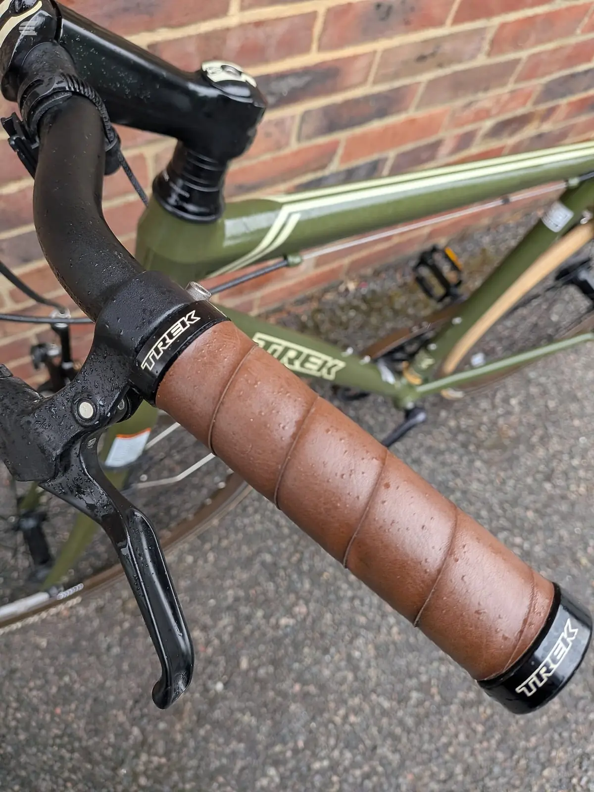 Close-up of Trek drop bar road bike handlebars with brown leather tape and black stem, against brick wall background.