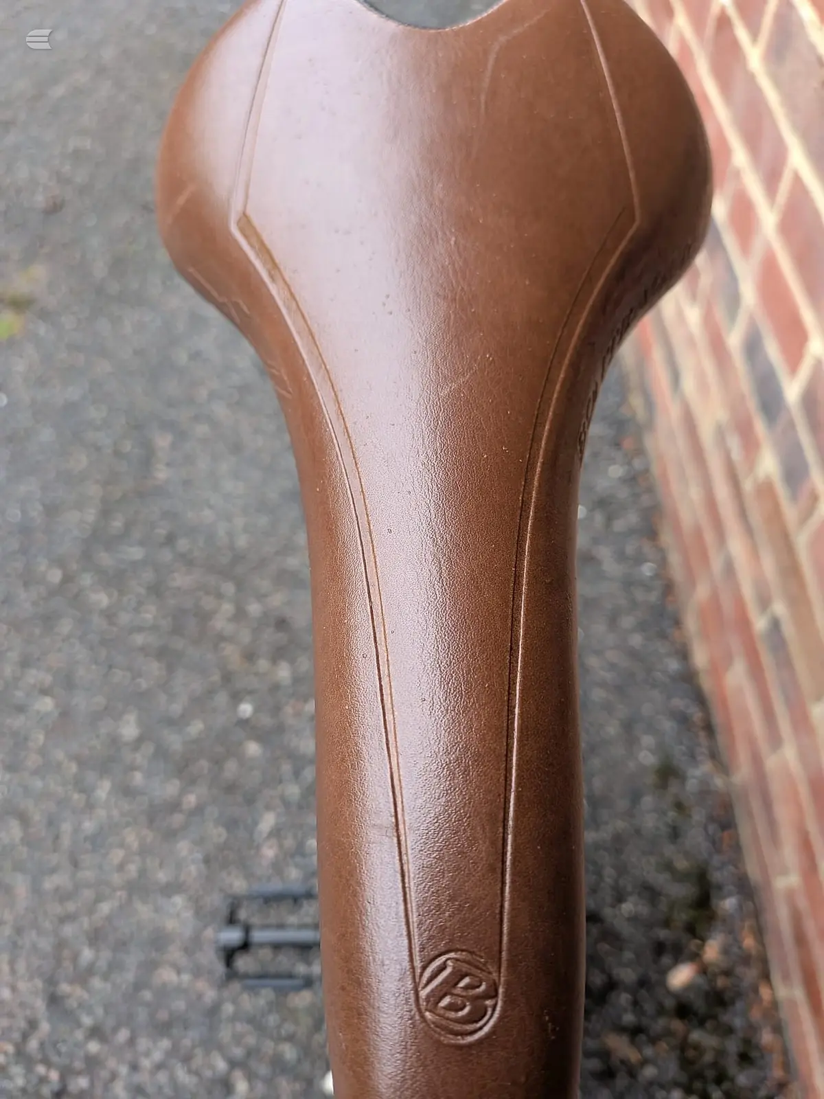 Brown leather saddle with decorative stitching and embossed logo, mounted on seatpost, against brick wall background.