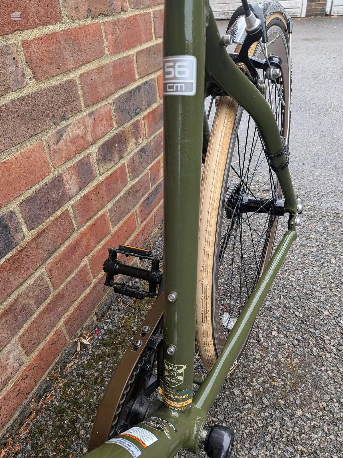 Olive-green road bike with wooden-rimmed wheels, leaned against red brick wall, non-drive side, parked on asphalt.