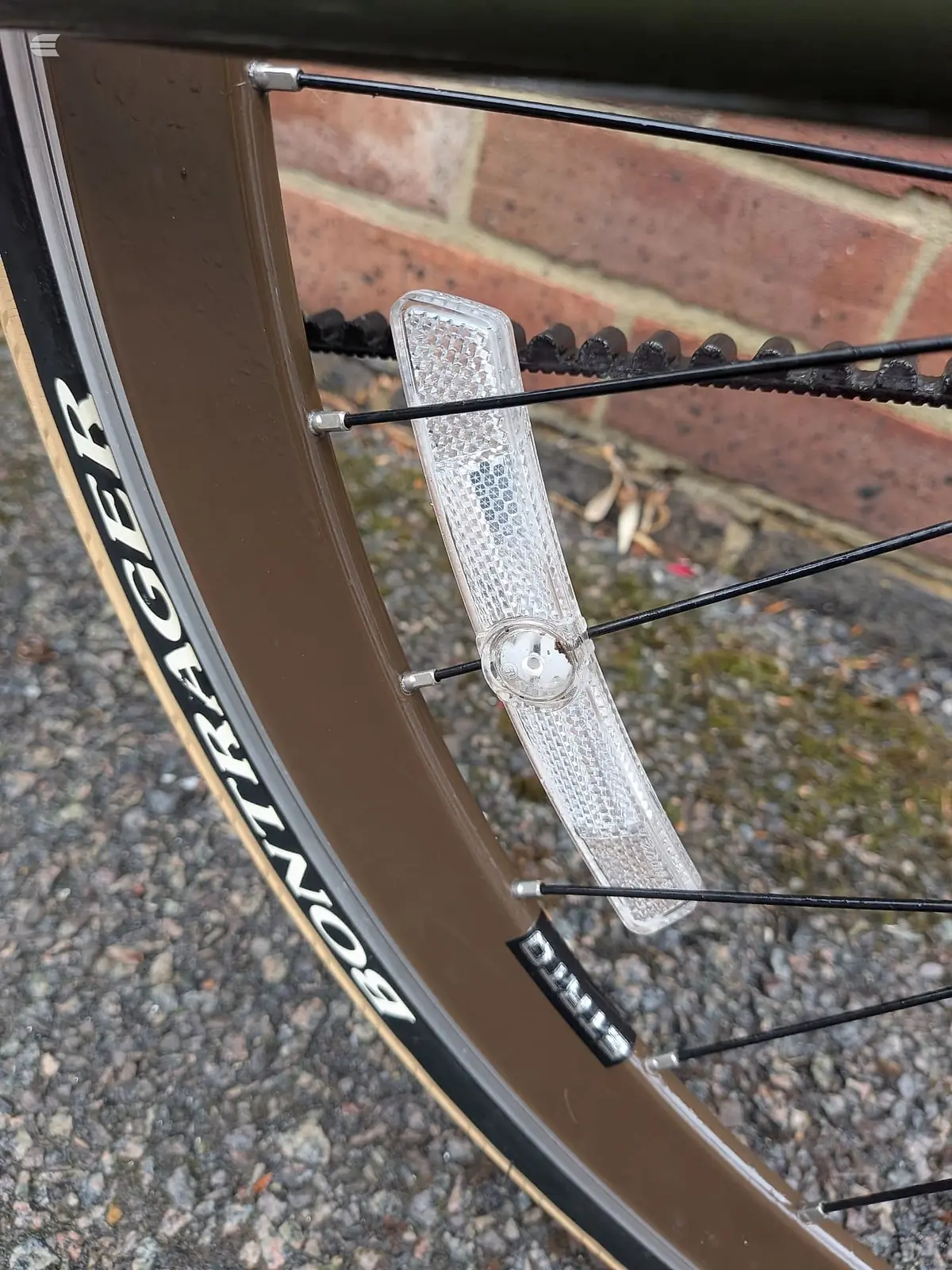 Close-up of a bicycle's reflector and brake cable assembly mounted on the frame, against a brick wall background.