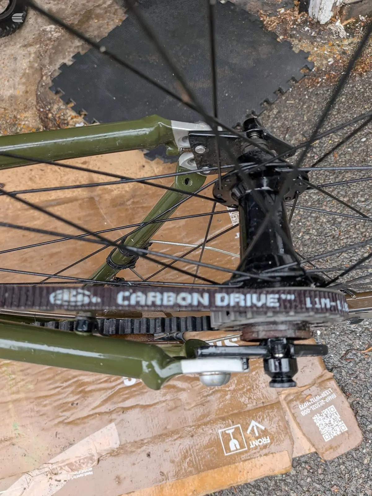 Olive-green bicycle frame with black disc brake and drivetrain components visible, drive side, on ground with cardboard packaging and black mat.