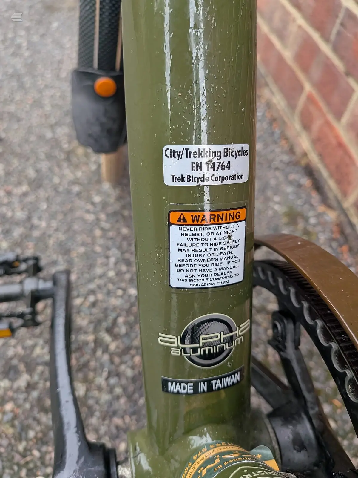 Close-up of olive-green aluminium bicycle frame tube with Trek City/Trekking Bicycles EN 14764 certification label and warning sticker, against brick