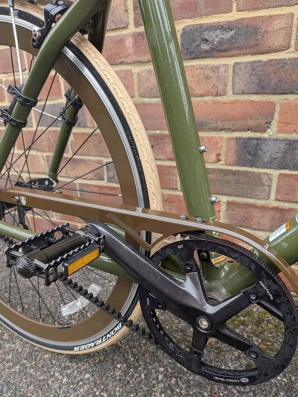 Green steel road bike, drive side close-up against brick wall. Focus on pedals, crankset, and bottom bracket area with chain and chainring visible.