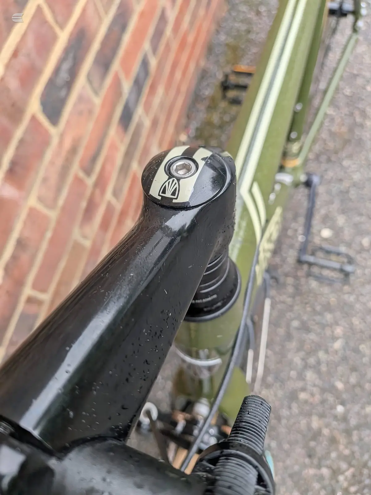 Close-up of black bicycle handlebar stem with integrated compass rose design, mounted on drop bars, against brick wall and green drainpipe.