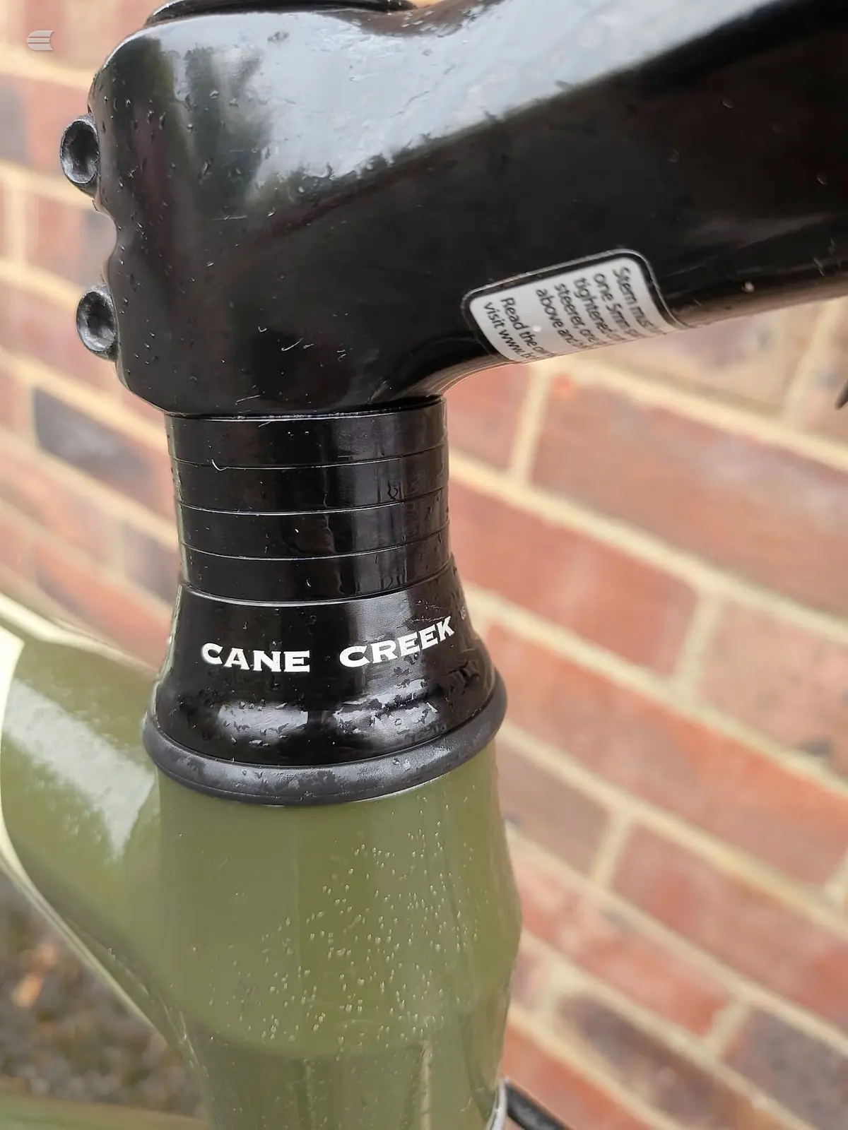 Close-up of Cane Creek bicycle stem and headset on green fork, mounted against red brick wall.