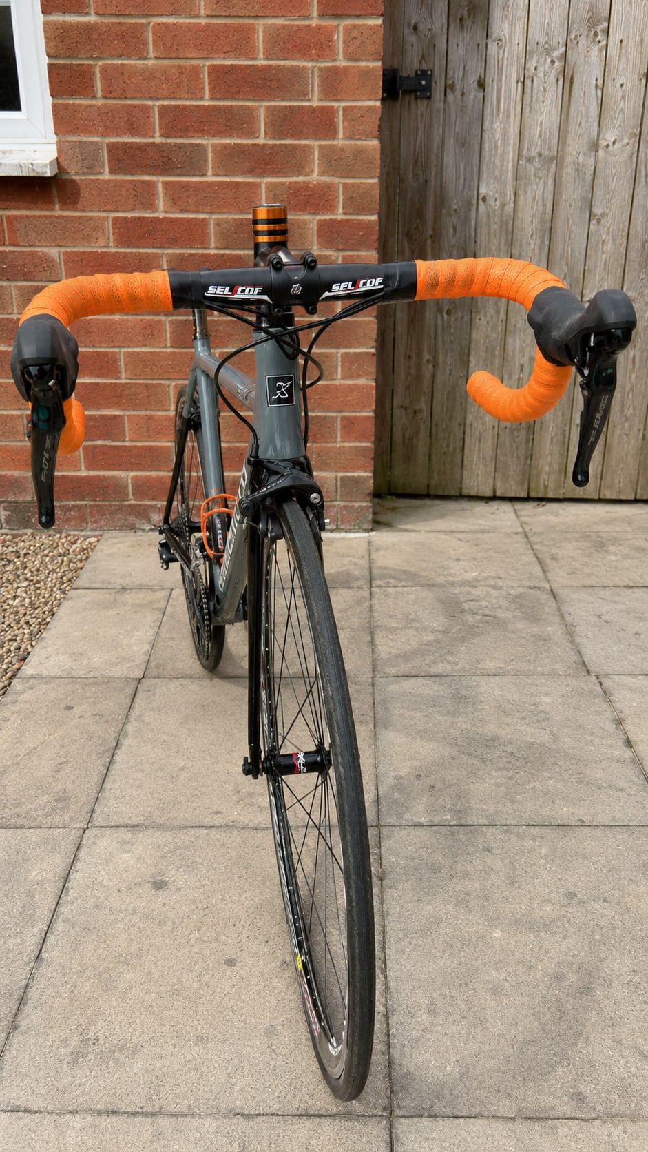 Grey and orange Selcof road bike, front-on view, on concrete paving against a brick wall and wooden fence.