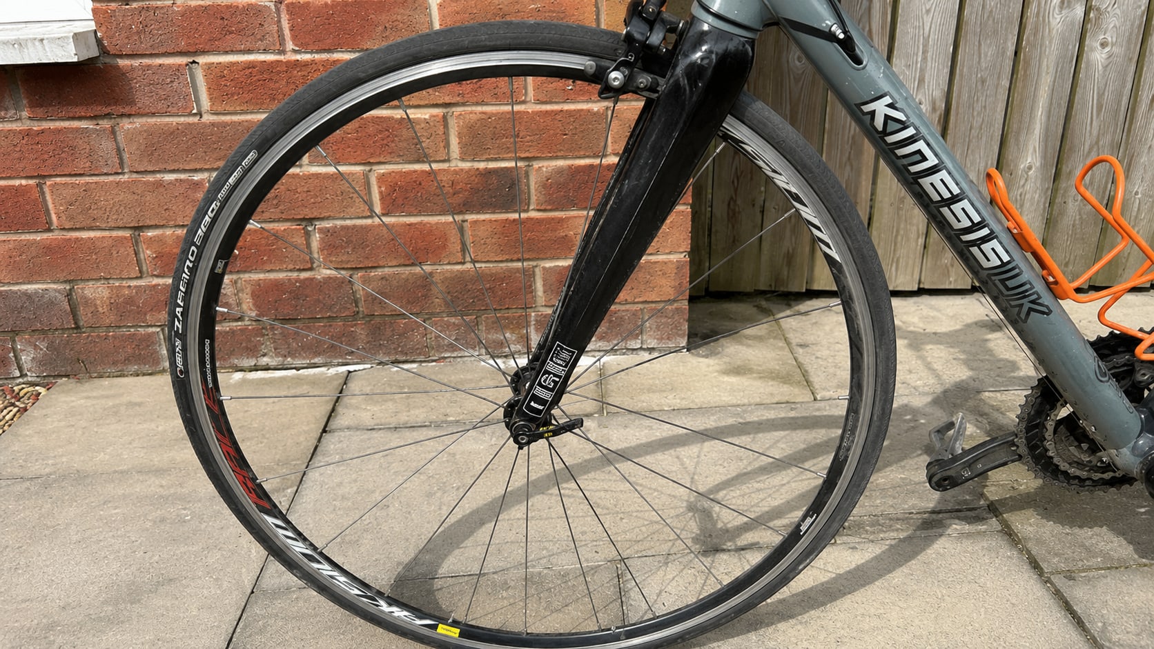 Black Kimberk road bike front wheel with Schwalbe tyre, non-drive side, leaning against red brick wall and wooden fence.